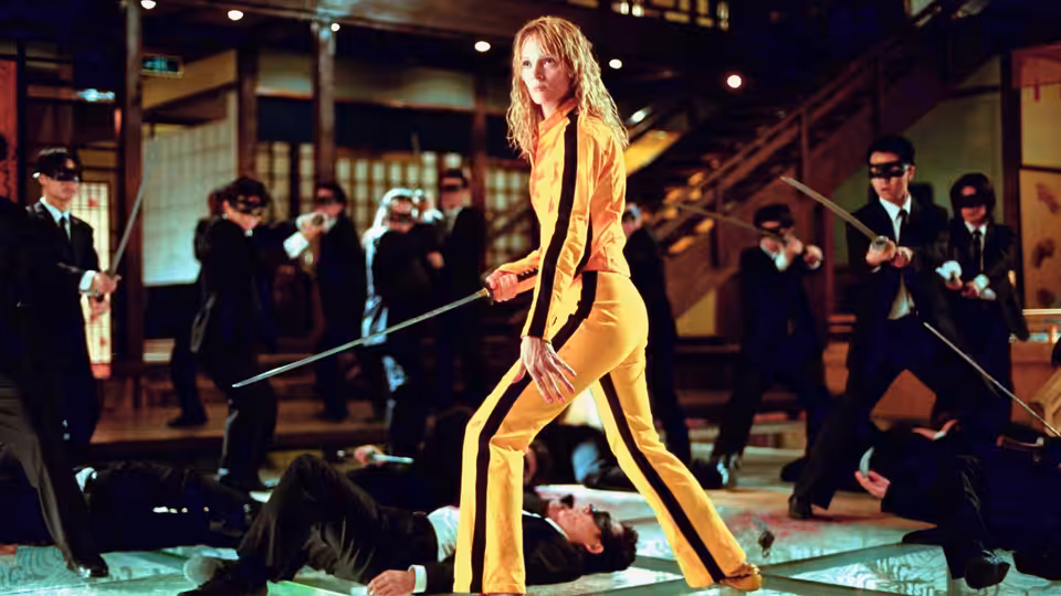 The Bride (Uma Thurman) in yellow tracksuit holds sword over fallen opponents