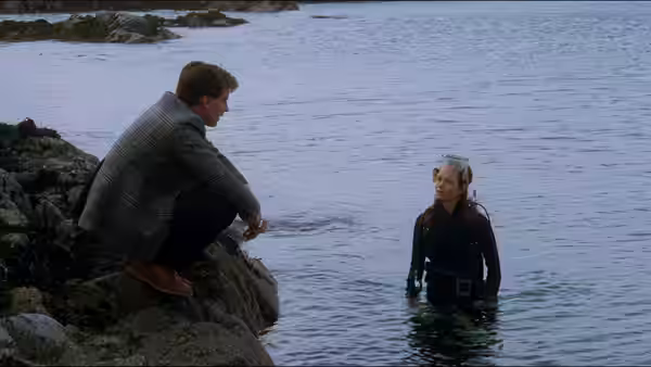 Mac (Peter Riegert) crouching on rocks speaking to Marina (Jenny Seagrove) in the water