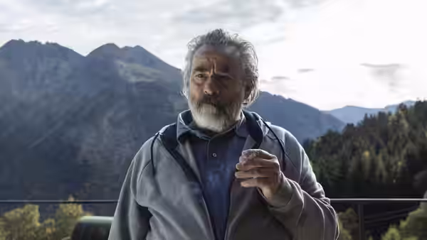 A grey-bearded man gestures outdoors before a forested mountain range