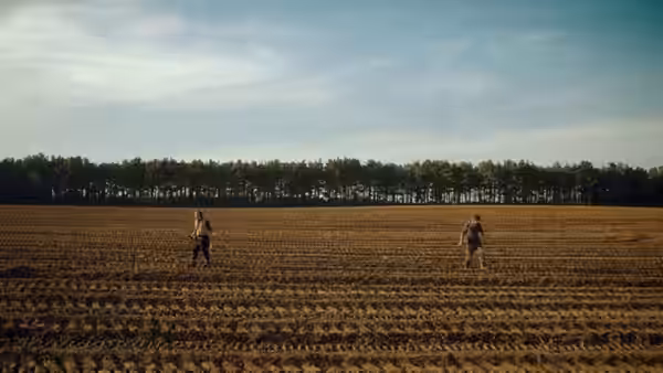 Two men walking across a vast plowed field toward a distant tree line