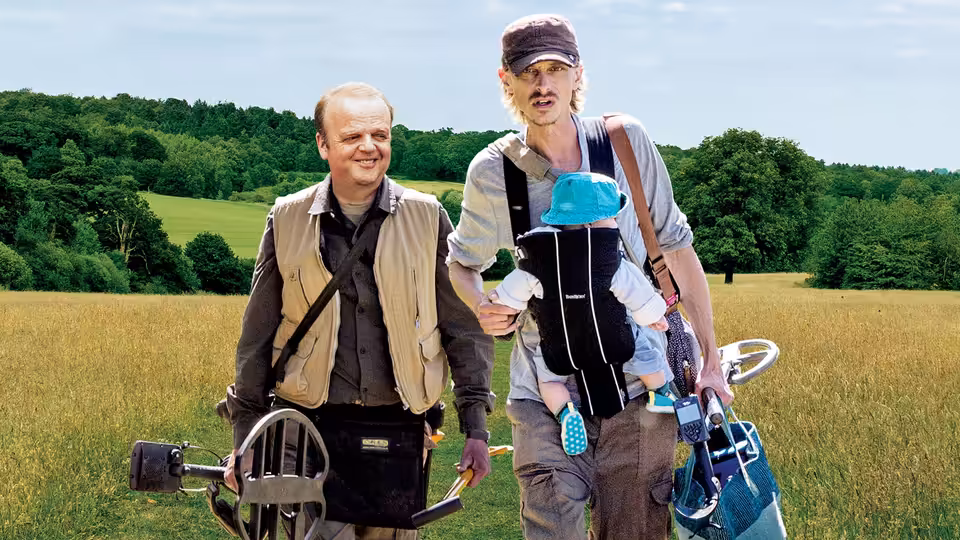 Lance (Toby Jones) and Andy (Mackenzie Crook) walking in a field with a baby