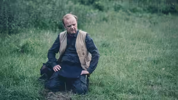 Lance (Toby Jones) kneeling in tall grass with a concerned expression