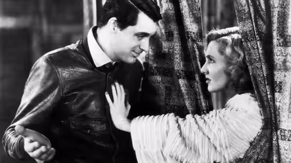 A man in a leather jacket leans toward a woman beside patterned curtains