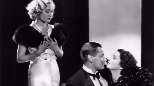 A woman stands behind Gaston and Lily as they kiss on a sofa