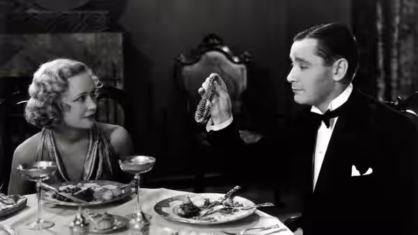 Mariette (Kay Francis) watches Gaston (Herbert Marshall) display a piece of jewelry over dinner