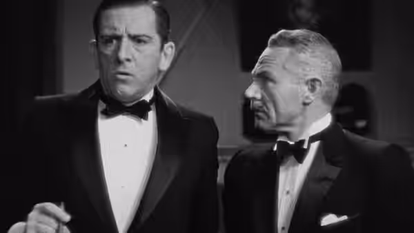 Two tuxedoed men stand together, one looking ahead while the other watches him