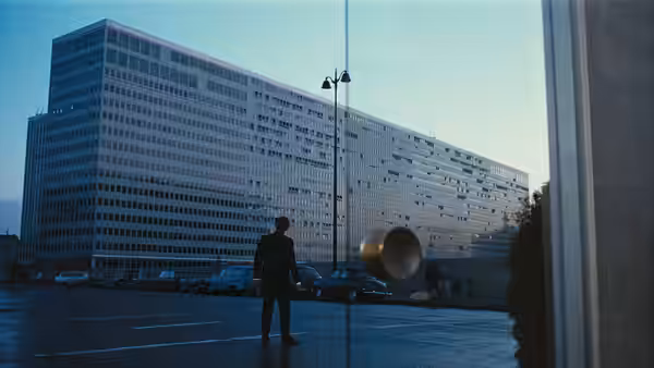 A solitary figure stands before a vast modern office building