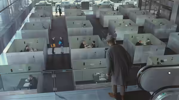 A man in a long coat walks through a maze of office cubicles