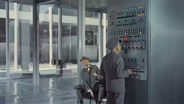 Monsieur Hulot (Jacques Tati) sits beside a man at a wall of control panels