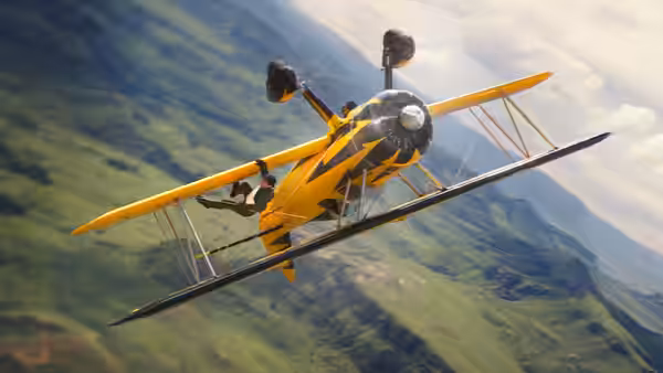 A man clinging to a yellow biplane flying over green mountains