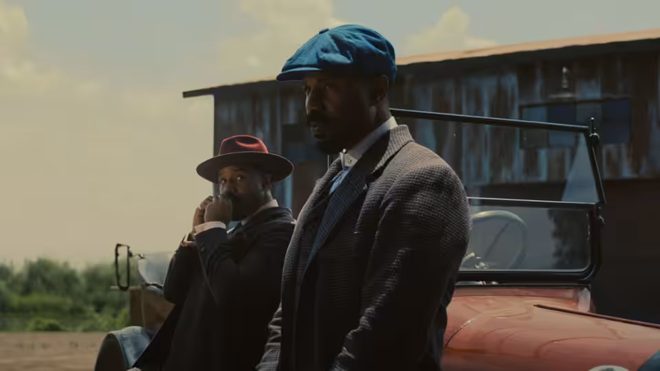 Michael B. Jordan as Smoke standing by a red car outside a wooden building with another man