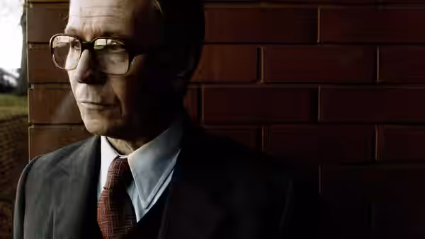 John Hurt as Control standing beside a red brick wall in a suit and tie