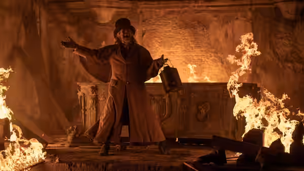 A man in a top hat with arms raised amid flames in a ruined hall