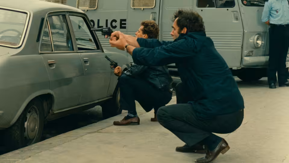 Two men crouch beside a parked car as police officers stand nearby