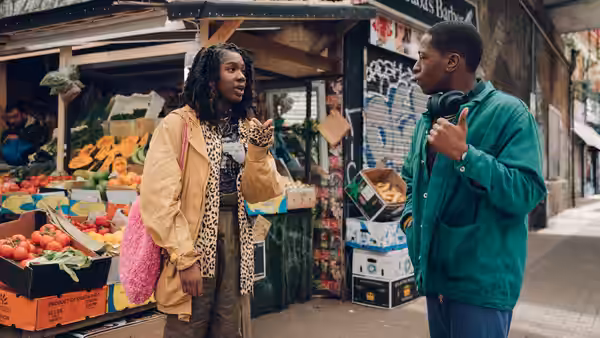 Yas (Vivian Oparah) and Dom (David Jonsson) talking outside a fruit and vegetable market stall