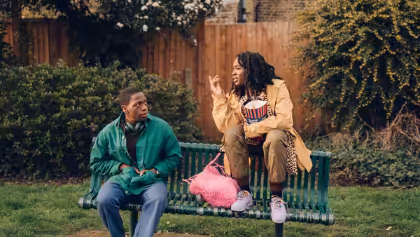 Dom (David Jonsson) and Yas (Vivian Oparah) sitting on a park bench eating popcorn