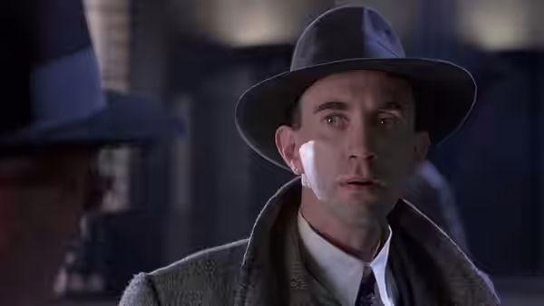 Man in fedora and overcoat with white cheek bandage facing another figure