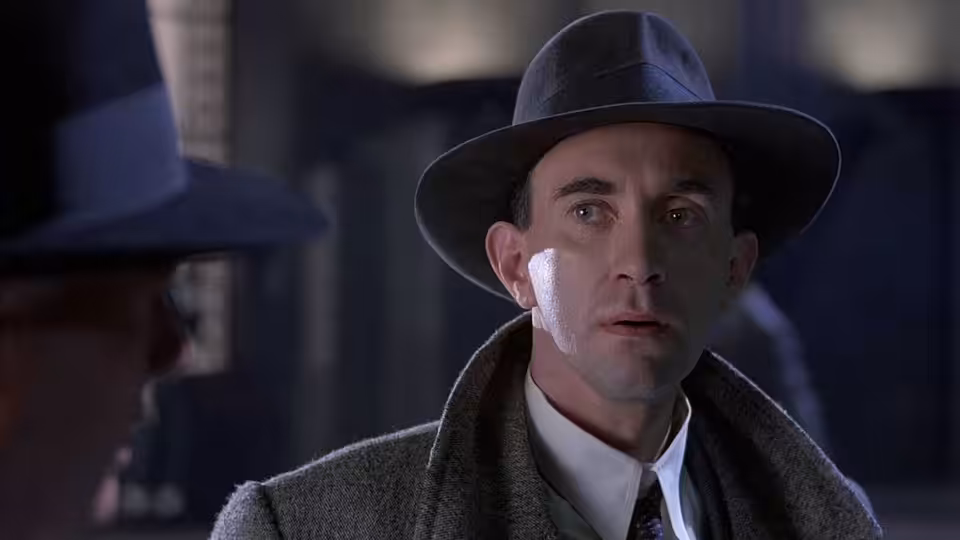 Man in fedora and overcoat with white cheek bandage facing another figure