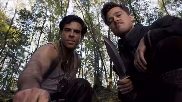 Two soldiers seated in a forest, one gripping a large knife