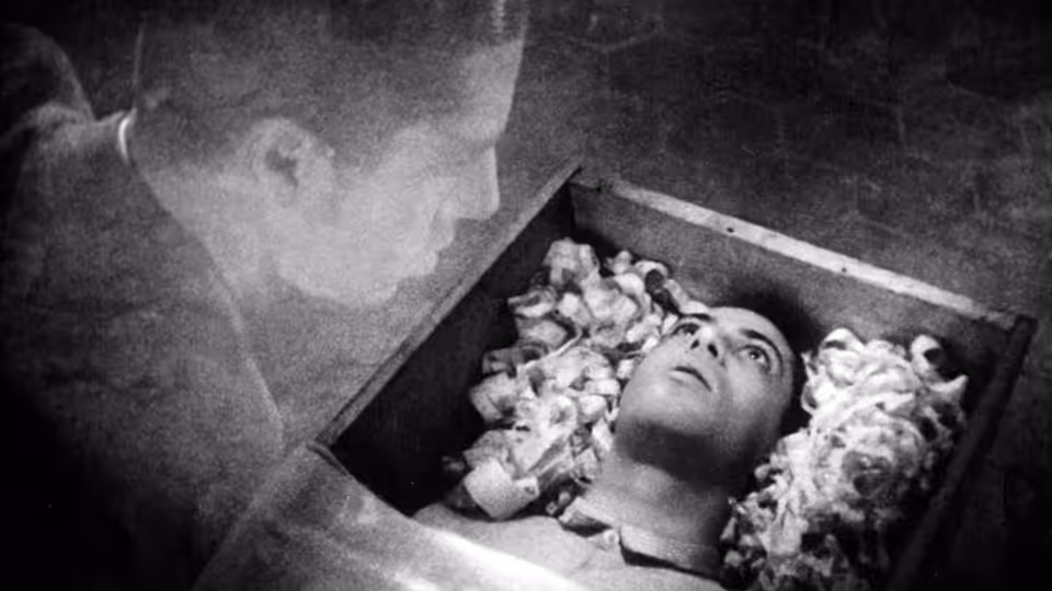 Allan Grey (Nicolas de Gunzburg) looks down at a body lying in a flower-lined coffin