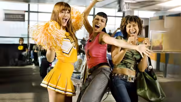 Three women cheering with arms raised in an airport terminal, one in cheerleader outfit