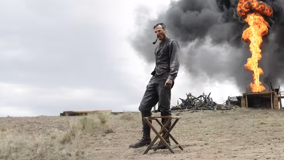 Daniel Plainview (Daniel Day-Lewis) stands by stool with huge fire behind him