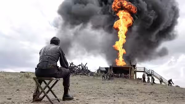 Man in leather jacket sits on folding chair watching large fire and explosion