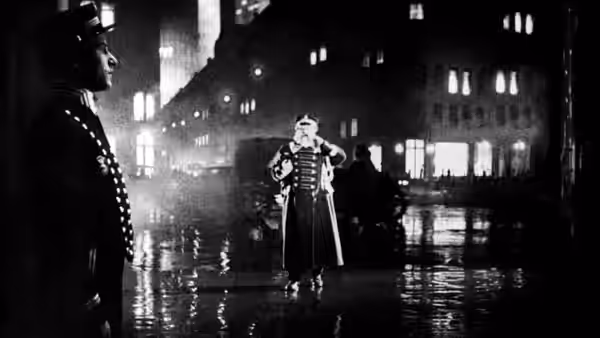 Hotel doorman Emil Jannings adjusts his uniform on a wet nighttime street as a man watches