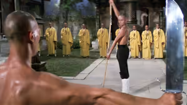A Shaolin monk faces a trainee wielding a broad blade in the courtyard