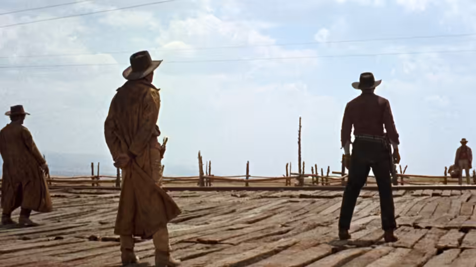 Four men in hats and dusters stand on a wooden platform in open country