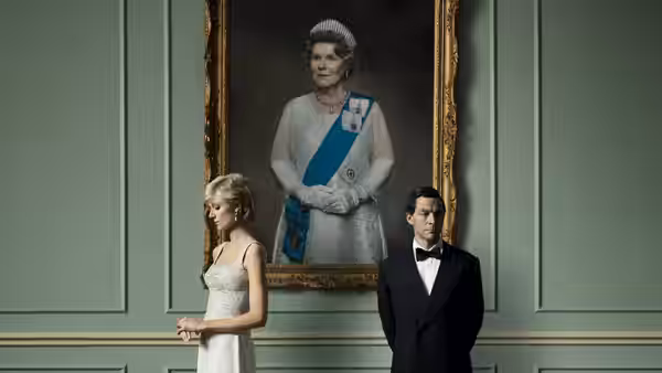 A woman in a white gown stands beside a man in a tuxedo before a portrait of Queen Elizabeth II