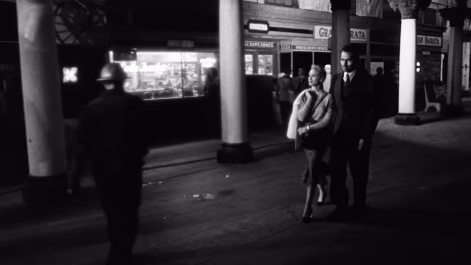 A woman and a suited man stand in a dim lobby as a shadowed figure approaches