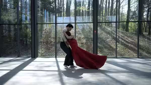A man supports a woman in a long red dress as they dance inside a glass pavilion