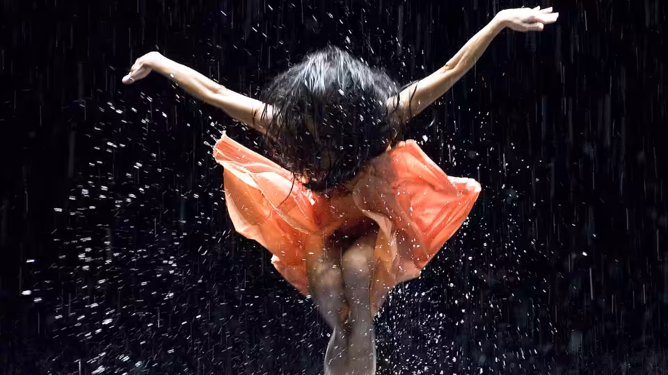 A dancer in an orange dress spreads her arms as water falls around her