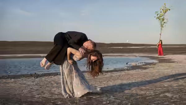 A woman bends beneath a man while another dancer stands beside a tree by the water