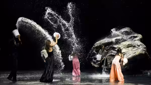 Dancers splash water across a dark stage beside a large artificial rock