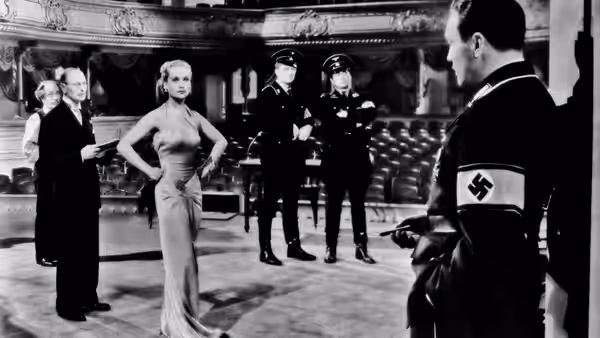 blonde woman in gown stands with hands on hips before uniformed men
