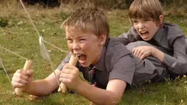 Will Proudfoot (Bill Milner) and Lee Carter (Will Poulter) shout and play with homemade props in the grass