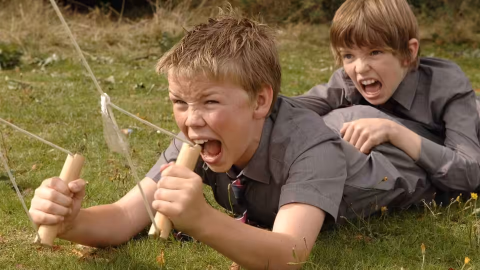 Will Proudfoot (Bill Milner) and Lee Carter (Will Poulter) shout and play with homemade props in the grass