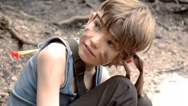 Will Proudfoot (Bill Milner) crouches on rocky ground with face paint and a toy dart tucked into his bag