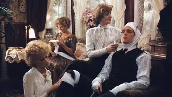 Women tend to a bandaged man seated in an armchair in a drawing room
