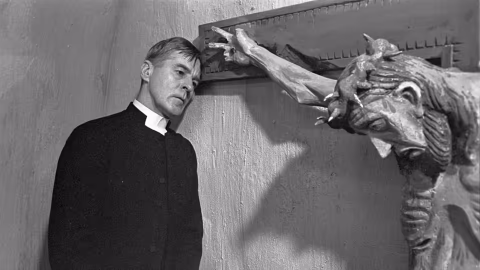 Tomas Ericsson (Gunnar Björnstrand) stands beside a large crucifix in the church