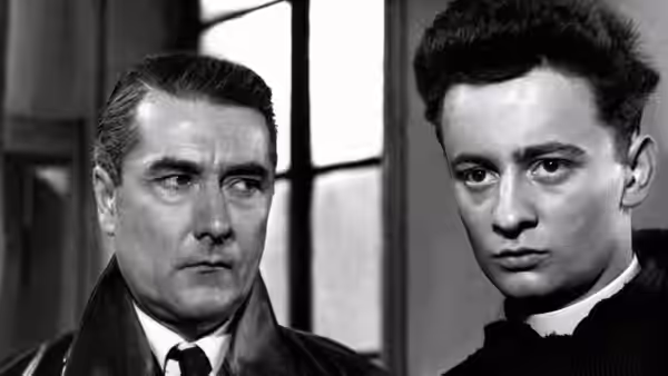 A priest stands beside a serious-looking man in a coat indoors