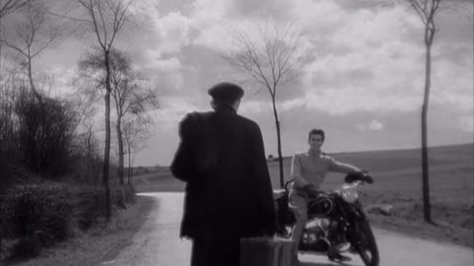 A priest stands beside a country road as a young man waits on a motorcycle