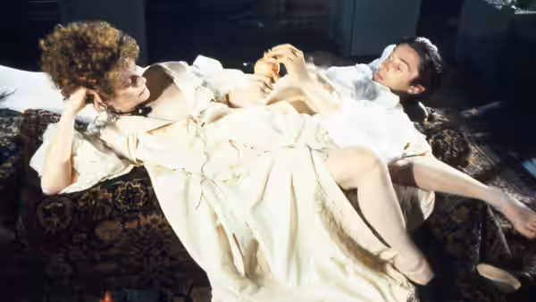 A woman in a pale gown reclines on cushions eating an apple near a man
