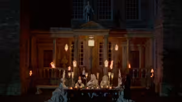 Period-dressed figures gather around a long table lit by candles outside a mansion