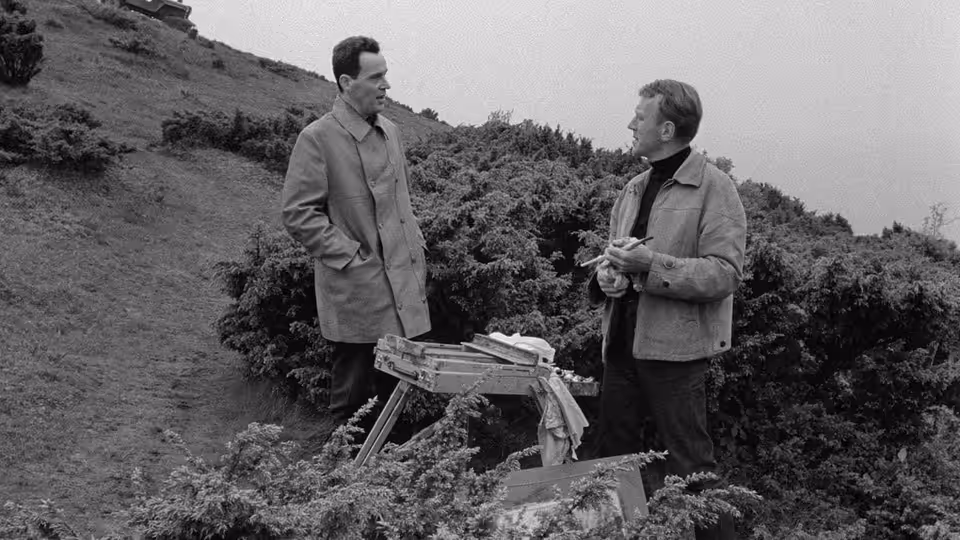 Two men talk beside an easel on a hillside