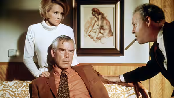 A woman stands behind a seated man as another man leans toward him