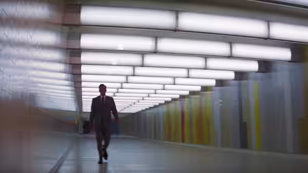 A suited man walks alone through a long corridor beneath rows of ceiling lights