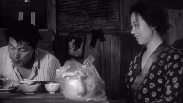 A man eats at a low table while a woman in a patterned robe smiles